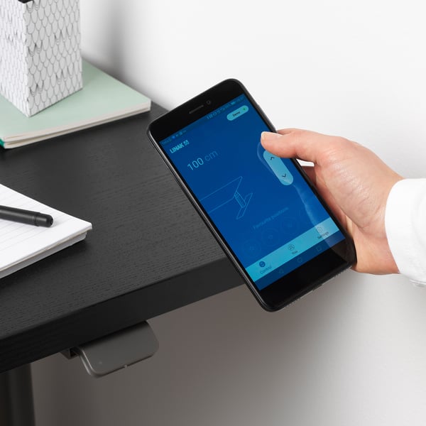 L’image montre une personne ajustant un bureau réglable en hauteur à l’aide de l’application de contrôle de bureau sur smartphone.