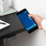 L’image montre une personne ajustant un bureau réglable en hauteur à l’aide de l’application de contrôle de bureau sur smartphone.