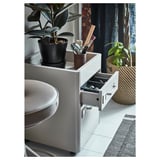 Un meuble à tiroirs IDÅSEN gris avec une plante en pot et des articles de toilette sur le dessus, situé dans une salle de bains avec des rideaux sombres.