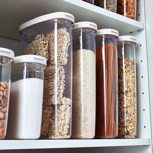 Les récipients de stockage transparents avec des couvercles blancs contiennent des aliments séchés comme les nouilles, le riz et l’avoine. Organisé sur une étagère blanche pour un accès facile.