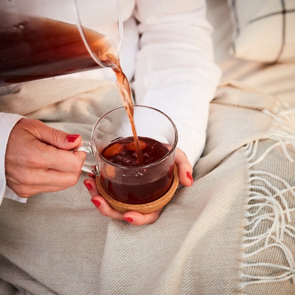 Une personne verse le café dans une tasse en verre sur un sous-verre en liège beige, protégeant la table de la chaleur et réduisant le bruit.