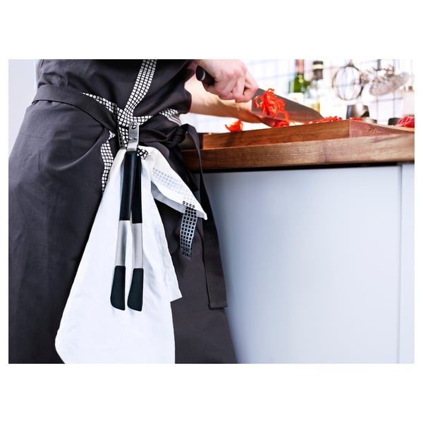 Personne tenant du poivron rouge avec une pince à épiler de cuisine, coupant sur une planche de bois avec un couteau de cuisine, portant un tablier noir et blanc.