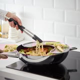 La personne utilise une pince à épiler pour cuire des nouilles et des légumes dans une casserole à plusieurs compartiments sur une cuisinière.