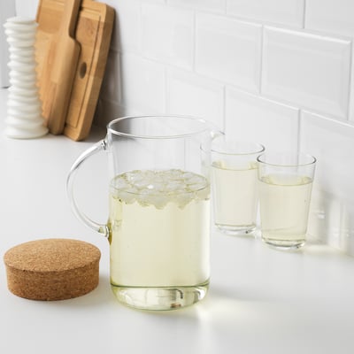 Pichet et tasses en verre transparent posés sur un comptoir blanc, remplis d’une boisson légère et de glaçons, accompagnés d’une planche en bois et de sous-verres.