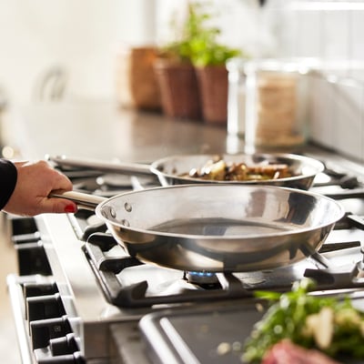 Une main tient une casserole en acier inoxydable sur une cuisinière à gaz. Une autre casserole avec des aliments en cuisson se trouve derrière, utilisant probablement la fonction de distribution uniforme de la chaleur des casseroles.