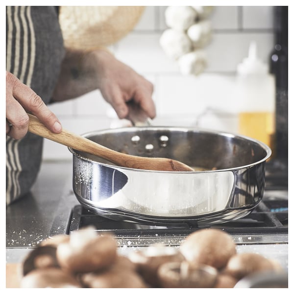 Une personne remue de la nourriture dans une casserole IKEA en argent sur une cuisinière. Des champignons se trouvent à proximité.