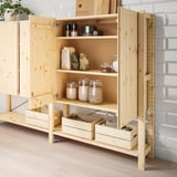 Étagère en bois avec portes ouvertes, présentant des pots de céréales et des sacs en papier, sur un sol en bois clair et un fond de mur en briques.
