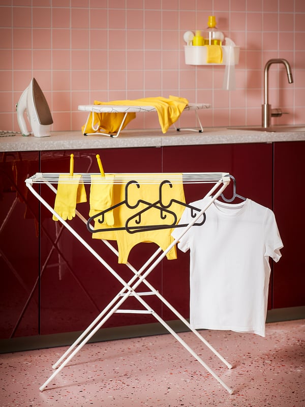 Une buanderie avec un étendoir JÄLL en blanc, contenant des vêtements jaunes et blancs, à côté d’un comptoir rouge et de murs carrelés de rose.