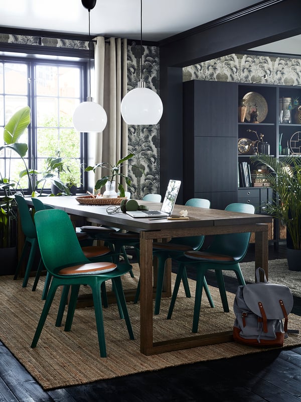 Salle à manger moderne avec table en bois, chaises vertes et deux suspensions globe blanches. Murs sombres, rideaux à motifs et plantes.