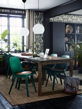 Salle à manger moderne avec table en bois, chaises vertes et deux suspensions globe blanches. Murs sombres, rideaux à motifs et plantes.