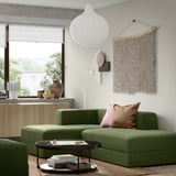 Salon moderne avec canapé vert JÄTTEBO, armoire en bois, suspension, art mural et table d’appoint avec livres et vase.