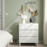 Chambre moderne avec commode blanche, lampe, miroir, plantes et crochets verts tenant des sacs à main. Des rideaux immatériels encadrent la scène.