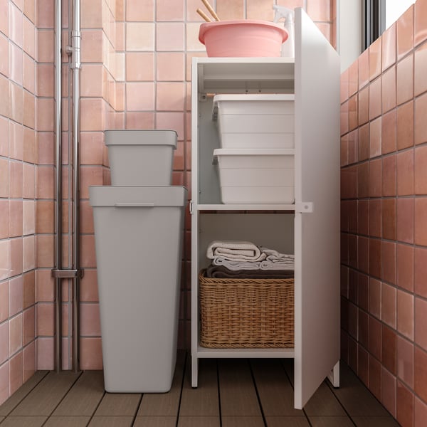 Petite armoire de rangement organisée dans la buanderie. Unité JOSTEIN blanche, panier en osier, piles de serviettes beiges. Poubelles grises, tuiles roses.