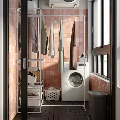 Buanderie compacte avec étendoir extensible JOSTEIN. Blanc, métal, murs carrelés, vêtements suspendus, laveuse et panier.