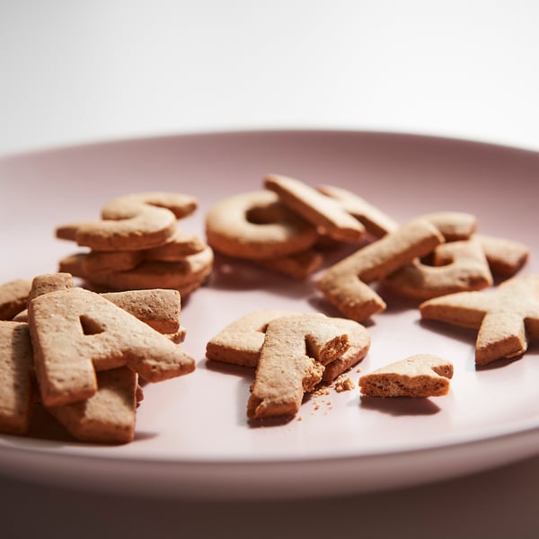 Des biscuits en forme de lettres brun clair épellent KAFFEREP sur une assiette rose.