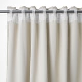 Les rideaux blancs pendent avec des plis, offrant de l’intimité et assombrissant la pièce. Languettes et crochets visibles en haut.