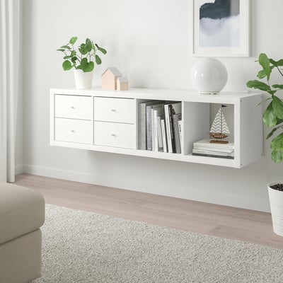 Des étagères blanches ikea KALLAX contiennent des livres, des plantes et des objets décoratifs, étroites avec des tiroirs, entre la décoration murale et un canapé.