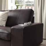 Description de l’image : canapé KIVIK gris foncé moderne avec un oreiller, placé à côté d’une fenêtre. Des écouteurs et une télécommande se trouvent au sommet du large accoudoir.