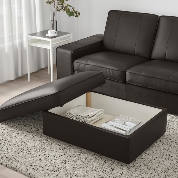 Canapé en cuir noir avec pouf ouvert, révélant des rangements contenant une couverture pliée et des magazines.
