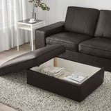 Canapé en cuir noir avec pouf ouvert, révélant des rangements contenant une couverture pliée et des magazines.
