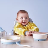 Bébé dans une chaise haute portant un bavoir à manches longues et une poche avant pour récupérer les aliments, conçu pour être confortable et facile à nettoyer.