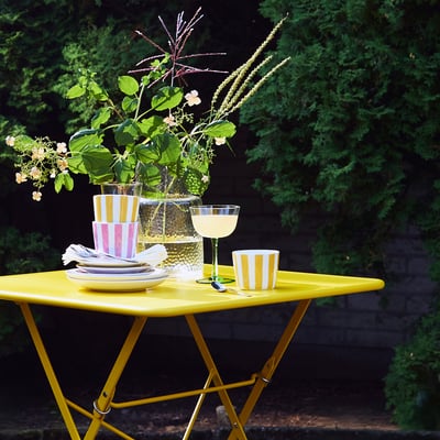 Ensemble de table d’extérieur jaune vif avec tasses et assiettes KLIPPAKLEJA, design rayé, porcelaine durable, entouré de verdure.