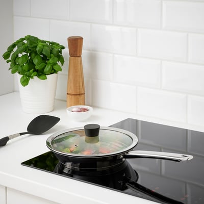 Scène de cuisson : poêle sur la cuisinière avec couvercle en verre, plante de basilic et moulin à poivre en bois.