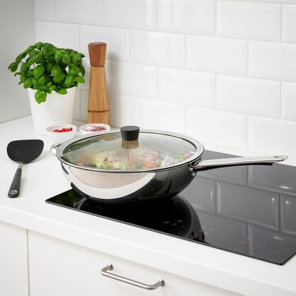 La poêle à frire en acier inoxydable avec couvercle en verre mijote les aliments sur une surface de cuisson noire près d’un mur de briques blanches.