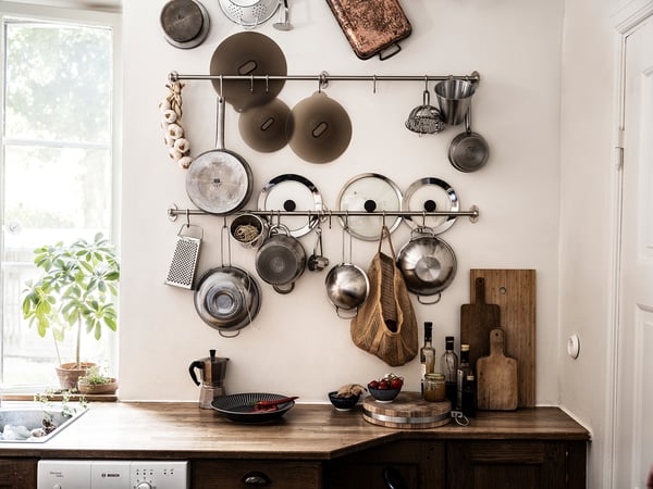 Mur de la cuisine avec des casseroles suspendues, des poêles et des couvercles organisés sur des étagères près d’une fenêtre. Comptoir en bois avec planches à découper, plantes et ustensiles en dessous.