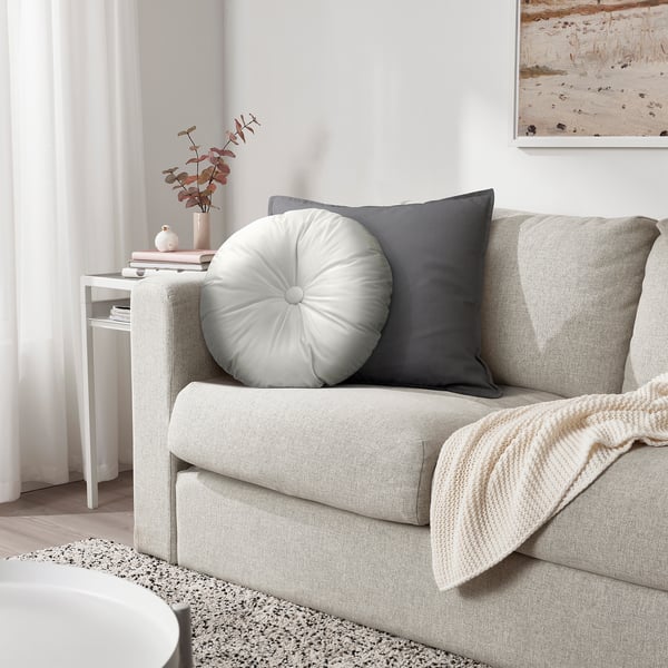 Salon moderne avec canapé texturé, oreiller en velours, coussin gris, couverture, table d’appoint avec vase et livres, impression de plage, éclairage chaleureux.