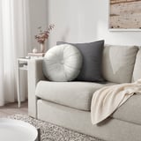 Salon moderne avec canapé texturé, oreiller en velours, coussin gris, couverture, table d’appoint avec vase et livres, impression de plage, éclairage chaleureux.
