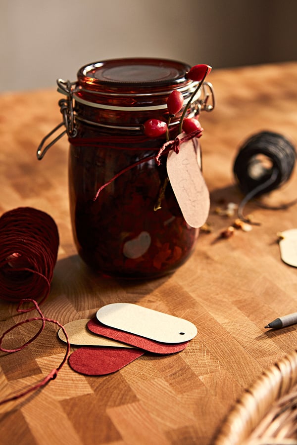 Pot en verre brun foncé avec des baies rouges, attaché avec de la ficelle et une étiquette, à côté d’étiquettes en bois, de fil et d’un stylo sur une table en bois.
