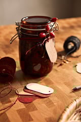 Pot en verre brun foncé avec des baies rouges, attaché avec de la ficelle et une étiquette, à côté d’étiquettes en bois, de fil et d’un stylo sur une table en bois.