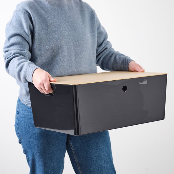 Personne tenant une boîte rectangulaire gris clair avec trou, portant un pull bleu et un jean.
