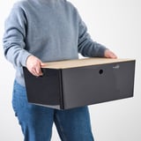 Personne tenant une boîte rectangulaire gris clair avec trou, portant un pull bleu et un jean.