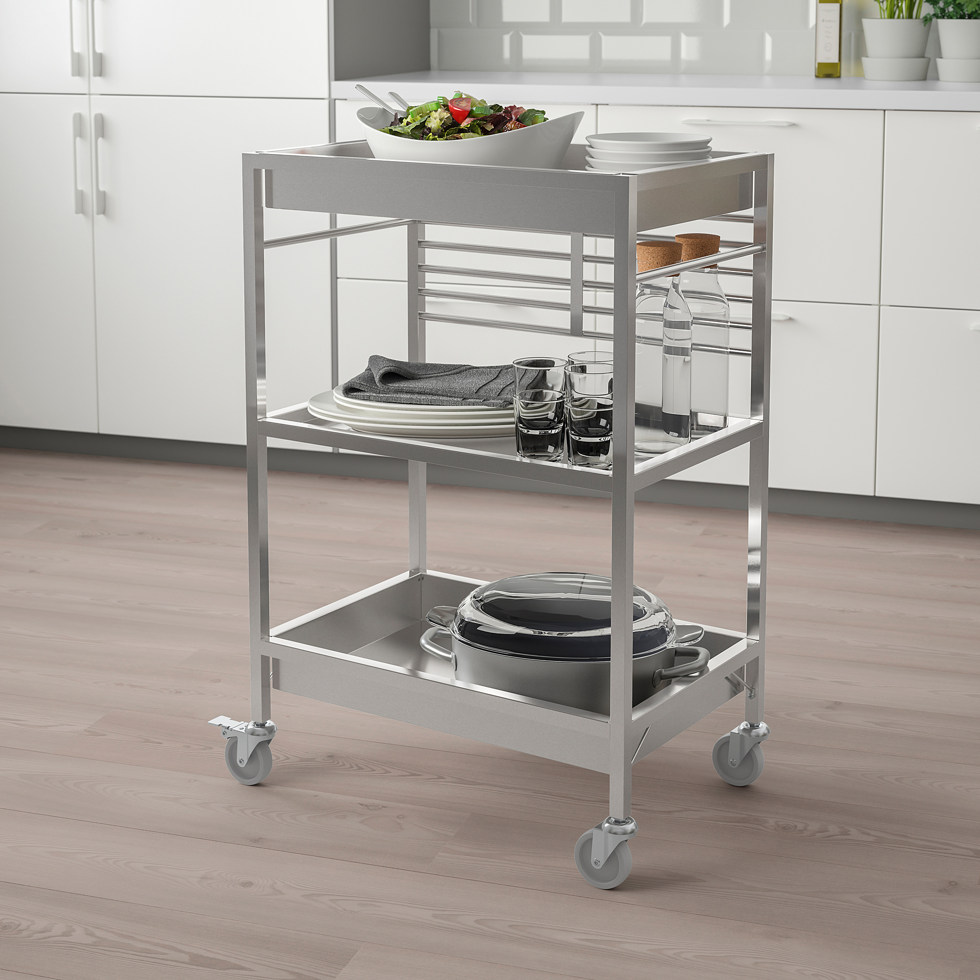 Chariot de cuisine KUNGSFORS argenté avec 3 étagères, des assiettes, des verres et des casseroles, s’intégrant parfaitement dans une cuisine moderne avec des armoires blanches.