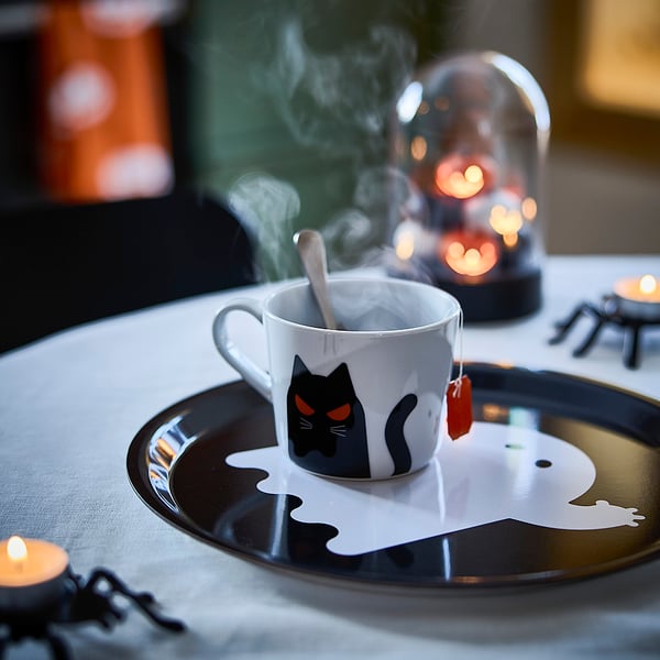 Table sur le thème d’Halloween avec tasse KUSTFYR , motif de chat noir, café fumant, cuillère et assiette ronde. Lumière de bougie en arrière-plan.
