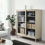 Armoire de rangement à porte vitrée KVARNVIK avec livres et décoration, à côté d’une chaise et d’une plante.