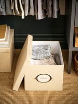 Une boîte de rangement KVARNVIK beige avec une étiquette ronde sur un sol noir, contenant des foulards pliés.