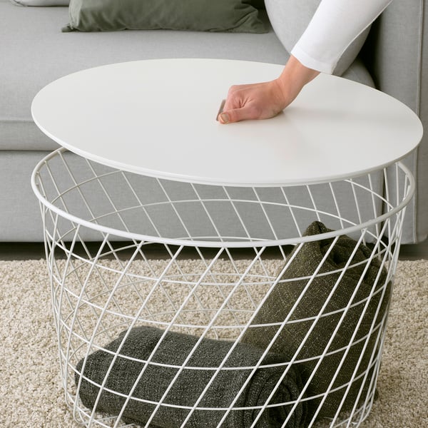 Personne soulevant une table KVISTBRO blanche avec un panier métallique. La table contient une couverture grise et un oreiller sur un tapis beige.