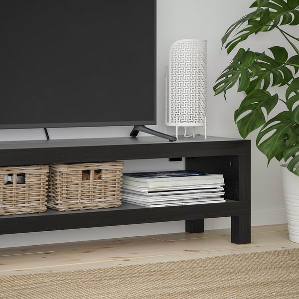 Meuble télé NOIR avec paniers tressés et livres empilés. Lampe blanche et grande plante verte à côté.