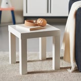 Petite table d’appoint LACK blanche avec plateau carré, quatre pieds groupés. Un livre et des accessoires beige se trouvent sur le dessus.