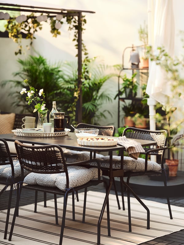 Ensemble de salle à manger d’extérieur avec chaises noires, coussins gris, table en bois, plantes et rideaux blancs.