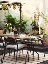 Ensemble de salle à manger d’extérieur avec chaises noires, coussins gris, table en bois, plantes et rideaux blancs.