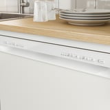 Cuisine moderne avec lave-vaisselle encastré blanc sous comptoir en bois. Panneau de commande visible, couverts et assiettes empilés sur le comptoir.