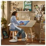 Espace de travail artistique : la femme est assise à une table LAGKAPTEN blanche avec un imprimé bleu, entourée d’un décor et de fournitures artistiques.