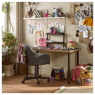 Salle d’artisanat avec bureau en bois, chaise de bureau noire, fil, projets de tricot, décoration murale, plantes et étagères présentant des articles colorés.