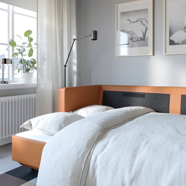 Chambre minimaliste avec lits jumeaux, literie blanche, tête de lit orange, applique murale, plante par fenêtre, art encadré, rideaux.
