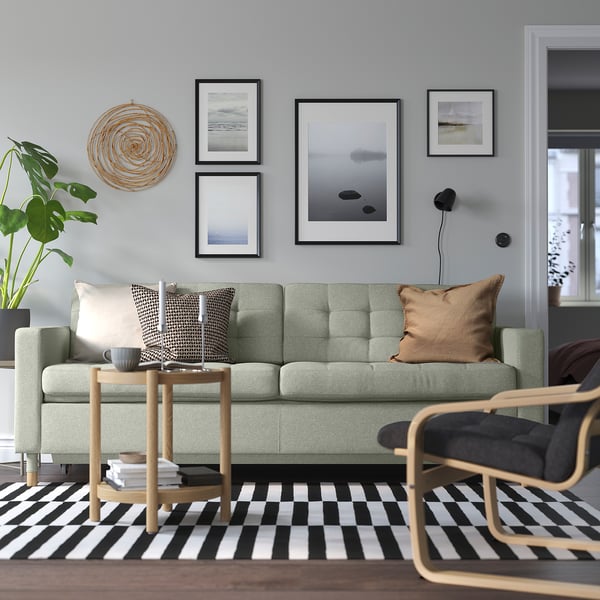 Salon moderne avec canapé vert LANDSKRONA. Table basse en chêne, tapis rayé et art mural. Le canapé est grand, avec des coussins et des oreillers.