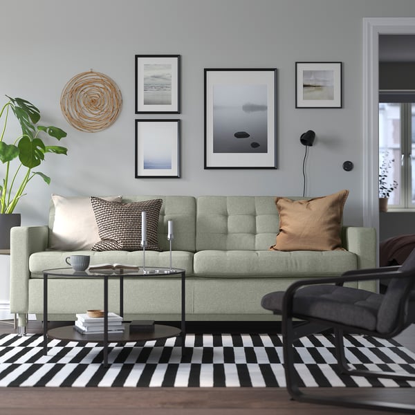 Salon moderne avec canapé LANDSKRONA vert, tapis noir et blanc, mur d’art et table basse avec des livres et une tasse.